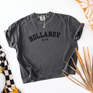 May include: A charcoal gray t-shirt with rolled-up sleeves. The shirt features the word "HOLLANOV" in black block letters, with "81-24" below it. A silver necklace with a pendant rests on the shirt.