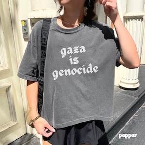 May include: A charcoal gray t-shirt with the words "gaza is genocide" printed in white. The shirt has a crew neck and short sleeves. The model is wearing a black skirt and a gold necklace.