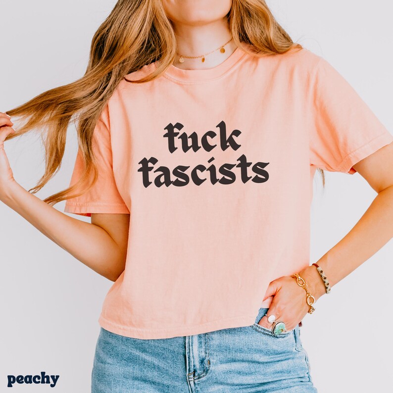 May include: Peach-colored t-shirt with the phrase "fuck fascists" in a bold, gothic font. The shirt is short-sleeved and has a relaxed fit. The word "peachy" is printed in the bottom left corner.