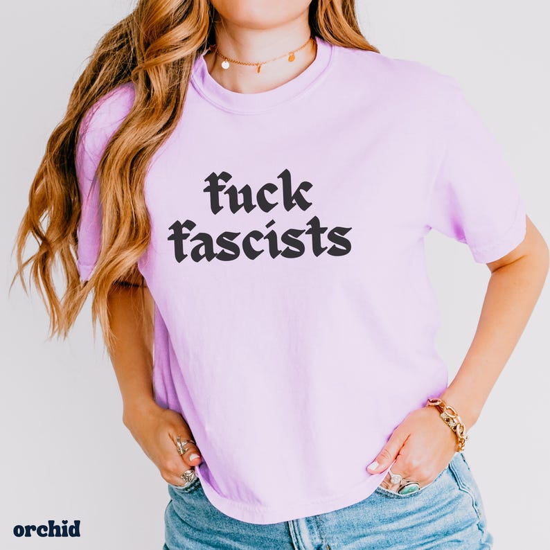 May include: A light purple t-shirt with the text "fuck fascists" in black gothic font. The word "orchid" is printed in the bottom left corner. The shirt is worn with blue jeans.