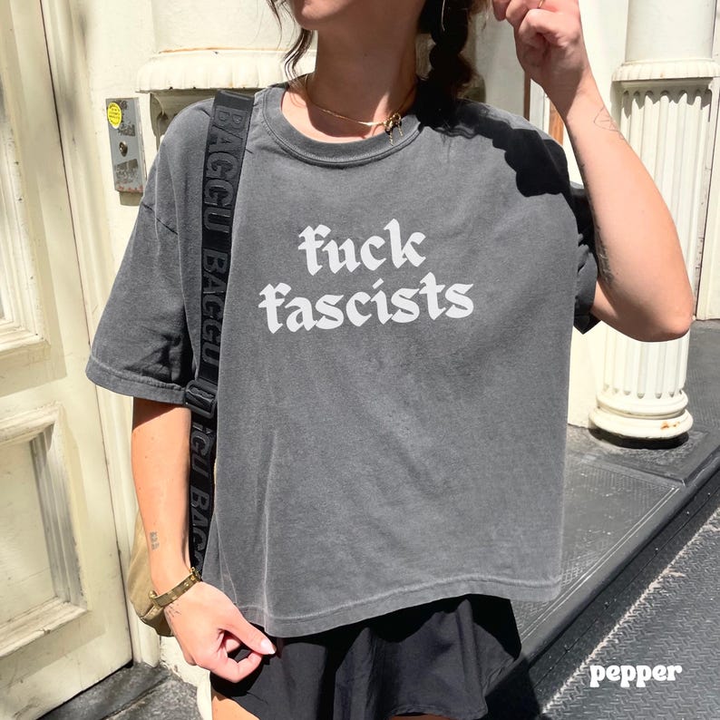 May include: A gray, short-sleeved t-shirt with the words "fuck fascists" in white, stylized lettering. The shirt is slightly faded, giving it a vintage look. The model is wearing a black skirt and a gold necklace.