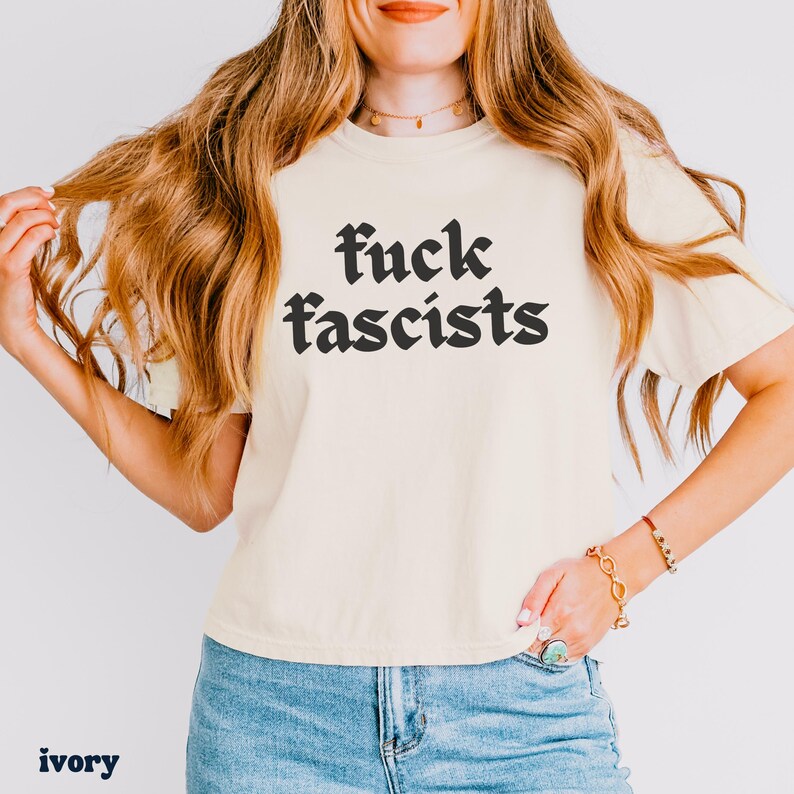 May include: Ivory-colored t-shirt with the words "fuck fascists" in a bold, black gothic font. The shirt is a short-sleeved crew neck. The word "ivory" is printed at the bottom left of the image.