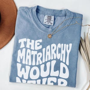 May include: A blue t-shirt with the text "THE MATRIARCHY WOULD NEVER" in white, retro-style lettering. A gold necklace with a pendant and a brown hat are also visible. The shirt is made of a soft material.