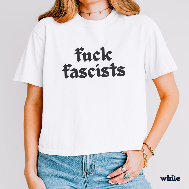 May include: White t-shirt with the phrase "fuck fascists" printed in black gothic lettering. The shirt is a crew neck style and is made of a soft material. The word "white" is printed in the bottom right corner.