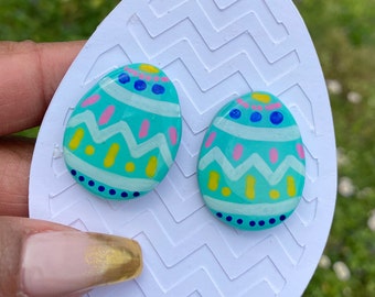 Könnte beinhalten: Ein Paar türkisblaue Osterei-Ohrringe mit weißen, rosa und gelben bemalten Designs.