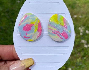Könnte beinhalten: Ein Paar bunte, ovale Ohrringe mit einem Marmormuster in den Farben Rosa, Blau, Gelb und Weiß. Die Ohrringe haben eine glitzernde Oberfläche.