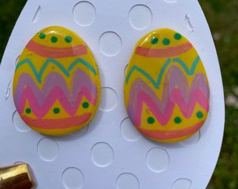 Könnte beinhalten: Ein Paar gelbe, rosa und grüne Ohrringe in Form von Ostereiern. Die Ohrringe sind mit einem geometrischen Muster bemalt und haben eine glänzende Oberfläche.