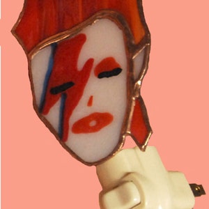 May include: A stained glass nightlight featuring a portrait of David Bowie with his signature lightning bolt makeup. The nightlight is attached to a white plastic base with an on/off switch.