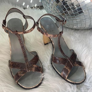 Peut inclure: Une paire de sandales à motif peau de serpent marron avec une bride en T, une bride de cheville réglable avec une boucle argentée et un talon bloc. La semelle intérieure est grise et porte le nom de la marque Gucci en lettres sombres.