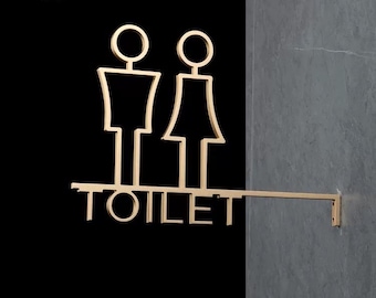 May include: A gold metal sign with the word "TOILET" and stylized male and female figures. The sign is mounted on a wall.