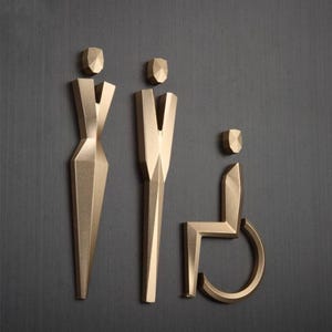 May include: Gold-colored metal restroom signs with a geometric design. The signs depict a person, a person in a wheelchair, and a person in a dress.