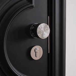 以下が含まれることがあります： 銀色のテクスチャード加工のドアノブと、お揃いの鍵穴カバーが付いた黒いドアのクローズアップ。 ドアノブは放射状のデザインで、鍵穴カバーは円形です。 ドアには装飾的な曲線パネルがあります。