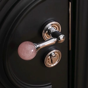 以下が含まれることがあります： 黒いドアのクローズアップ。磨かれたクロムとローズクォーツのドアハンドルが付いています。ドアハンドルは丸いローズクォーツのノブと黒い円筒形のハンドルが付いています。ドアにはクロムと黒のドアロックも付いています。
