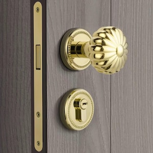 Könnte beinhalten: Ein goldfarbenes Türgriff-Set mit einem blütenförmigen Knauf und einer Schlüssellochabdeckung. Das Set enthält eine lange, rechteckige Platte für die Türkante. Die Hardware ist an einer Holztür montiert.