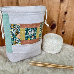 May include: A fabric drawstring project bag featuring a patchwork design with floral patterns, a cat illustration, and solid panels in light grey, green, and brown. Beside the bag sits a ball of off-white yarn and four wooden knitting needles.