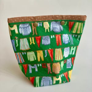 Può includere: Una borsa tote in tessuto verde con un motivo ripetuto di vestiti appesi a un filo per stendere. I vestiti sono di vari colori e stili, tra cui maglioni, pantaloni e gonne. Il bordo superiore è marrone con piccoli motivi a stella e due bottoni marroni.