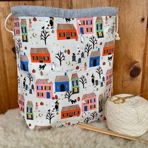 May include: A drawstring project bag featuring a cream-coloured fabric with a repeating Halloween pattern of colourful houses, trees, ghosts, and pumpkins. The bag has a grey fabric top edge and white cords. A ball of cream yarn and wooden needles sit nearby.