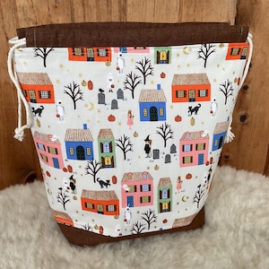 May include: A drawstring bag featuring a cream-coloured fabric with a repeating pattern of colourful houses, black trees, pumpkins, ghosts, and gravestones. The bag has a solid brown fabric top border and base, with white rope drawstrings on each side.