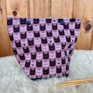 Può includere: Una borsa in tessuto per progetti con un motivo ripetuto di volti di gatto stilizzati in tonalità viola scuro, viola chiaro e nero. La borsa presenta un'ampia apertura fissata con due bottoni a pressione circolari scuri sul bordo superiore.