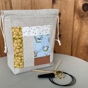 Op de afbeelding: Een lichtgrijze projecttas met trekkoord en een patchwork voorpaneel met stoffen vierkanten in bloemen-, effen- en ganzenpatronen. Naast de tas liggen twee houten breinaalden met een zwart koord en een goudkleurige schaar.