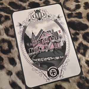 May include: A black and white illustration of a pink house with a red flag, framed by a decorative oval border with a beetle and button. The house has multiple stories and a detailed architectural design.