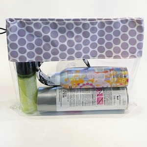 May include: A clear plastic zippered pouch with a purple and white polka dot fabric lining. The pouch contains three bottles of hair care products. The bottles are labeled "TRISH FRAGRANCE EXTRA AQUA NET ALL DAY HOLD" and "L'oreal Paris"