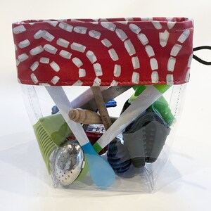 May include: A clear plastic bag with a red and white patterned fabric top. The bag is filled with various kitchen utensils, including a green silicone brush, a blue spoon, a wooden spoon, a green tea strainer, and a black oven mitt.