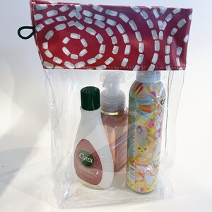 May include: Clear plastic zippered pouch with a red and white patterned top. The pouch contains three bottles of toiletries: a clear bottle with a green lid labeled "Cutex" and a pink bottle with a gold lid and a yellow bottle with a colorful design.