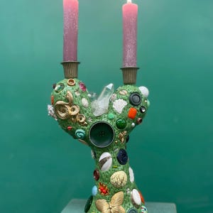 Puede incluir: Un candelabro verde con forma de cactus que sostiene dos velas cónicas rosas encendidas. La superficie está decorada con varios adornos, incluyendo una serpiente dorada, una mariposa, botones, cuentas y un cristal de cuarzo claro en el centro.