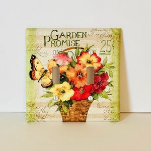 May include: A light switch cover with a floral design. The cover features a basket of flowers with butterflies and the text "Garden Promise".