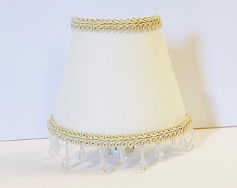 Può includere: Un paralume bianco con bordino dorato e frange di cristallo. Il paralume ha una forma conica e un design semplice.