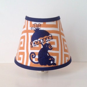 May include: A decorative night light shade featuring a geometric orange and white pattern. The shade has a navy blue trim and a navy blue embroidered monkey holding an umbrella. The night light is designed to plug into a wall outlet.