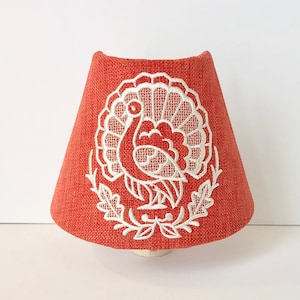 May include: A red lampshade with a white embroidered turkey design.