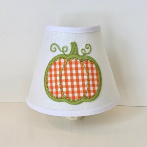 May include: A white nightlight lampshade with a green and orange pumpkin design. The pumpkin is made of gingham fabric with a white background and orange squares.