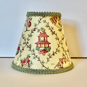 May include: A cream-colored lampshade with a green trim and a floral pattern featuring red pagodas and green vines.