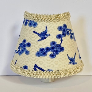 May include: A white nightlight lampshade with a blue and white floral pattern featuring birds. The lampshade has a cream-colored trim around the top and bottom.