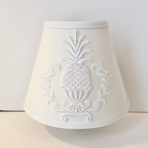May include: Off-white lampshade with a white embroidered pineapple design. The lampshade has a classic, tapered shape and is trimmed with white fabric along the top and bottom edges. The pineapple is surrounded by decorative floral elements.
