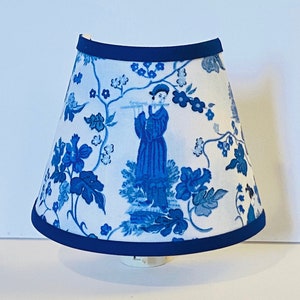 May include: A blue and white lampshade with a floral pattern and a figure playing a flute. The lampshade has a dark blue trim.