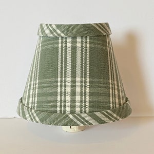 May include: A green and white plaid lampshade with a white trim. The lampshade is a cone shape and is designed for a nightlight.