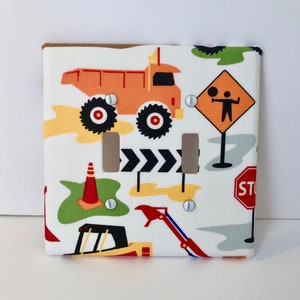 May include: A white fabric light switch cover featuring a construction theme. The design includes an orange dump truck, road signs, and construction vehicles. The cover has two light switch openings and is secured with silver screws.