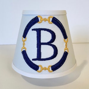 May include: White lampshade with a blue and gold embroidered design. The design features a circle with a large letter "B" in the center. The circle is surrounded by a blue band with gold accents.