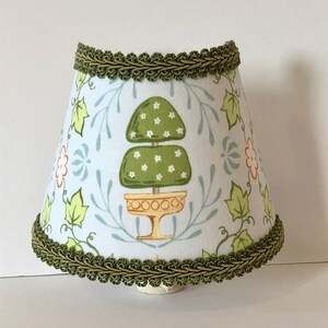 May include: A white lampshade with a green and white floral pattern. The lampshade has a green trim around the top and bottom.