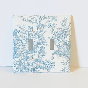 May include: A white light switch cover with a blue and white toile pattern featuring a pastoral scene. The switch cover has two toggle switches.