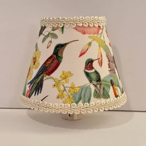 May include: A white lampshade with a floral pattern featuring hummingbirds and flowers. The lampshade has a white braided trim around the top and bottom.