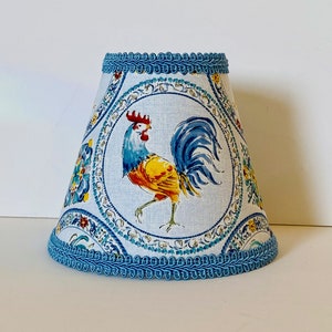 May include: A blue and white lampshade with a rooster design. The rooster is yellow, red, and blue. The lampshade has a blue trim around the top and bottom.