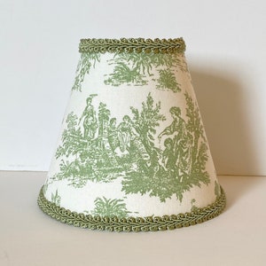 May include: A green and white lampshade with a toile pattern and green trim. The lampshade is cone-shaped and has a scalloped edge.