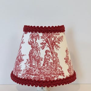 May include: A red and white toile lampshade with a red braided trim. The lampshade features a scene of a woman and two children in a garden.