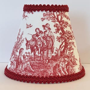 May include: A red and white toile lampshade with a red braided trim. The lampshade features a pastoral scene with people and animals.