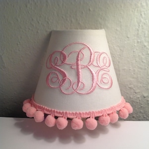 May include: A white lampshade with a pink monogram and a pink pom-pom trim.