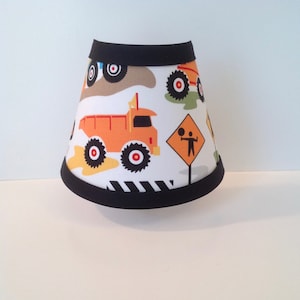 May include: A white lampshade with a black trim features a construction theme with orange dump trucks, black and white striped tires, and a yellow road sign with a stick figure.